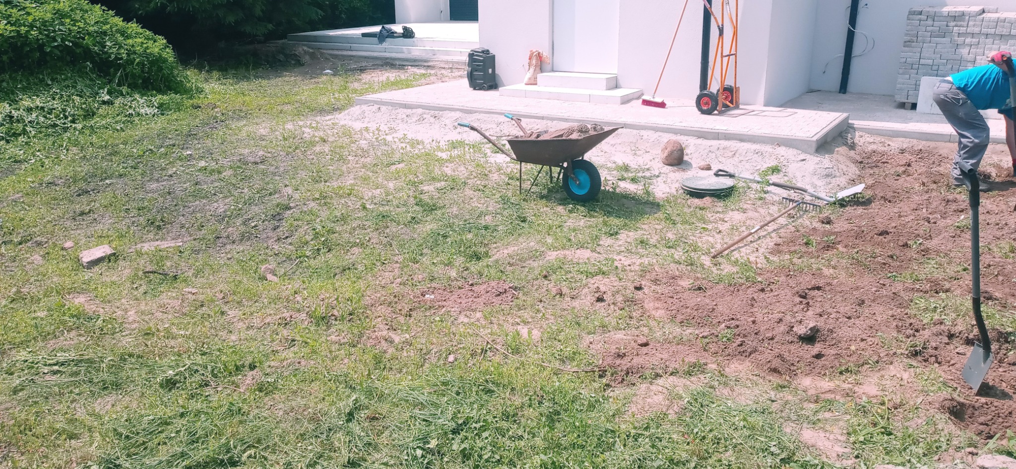 Przygotowanie terenu pod ogród: pracownik z łopatą kopie ziemię, obok taczka z ziemią, grabie i paleta z kostką brukową w tle.