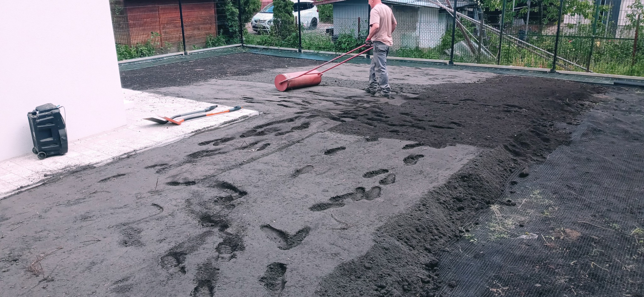 Mężczyzna wyrównuje teren wałem ogrodowym, widoczne ślady stóp na świeżo rozplantowanej ziemi, obok ściany budynku leżą dwie szpadle, w tle siatka ogrodzeniowa i zabudowania.