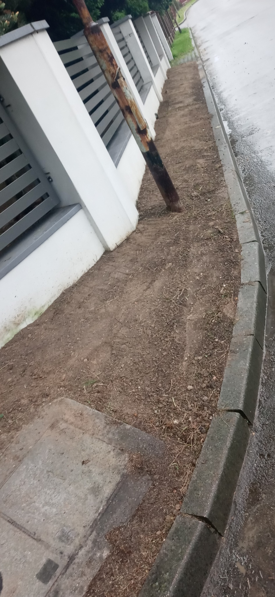 Przygotowany teren pod trawnik obok białego ogrodzenia z widocznym rdzewiejącym słupkiem i betonowym obrzeżem chodnika.