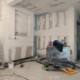 RAJ-BUD Usługi Ogólnobudowlane Robert Fujak - Wnętrze w trakcie prac wykończeniowych: ściany i sufit pokryte gładzią, widoczne przewody elektryczne, narzędzia malarskie i meble zabezpieczone folią.