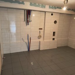 RAJ-BUD Usługi Ogólnobudowlane Robert Fujak - Wnętrze łazienki w trakcie remontu, widoczne białe płytki ścienne z szarą fugą, szare płytki podłogowe, wystające rury instalacji wodnej i elektrycznej, oraz fragment ściany z nałożoną gładzią...