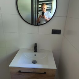 RAJ-BUD Usługi Ogólnobudowlane Robert Fujak - Nowoczesna łazienka z białą umywalką nablatową na szafce z drewnianym fornirem i czarnymi uchwytami, czarna bateria umywalkowa, białe płytki na ścianach i okrągłe lustro z czarną ramą, w którym...