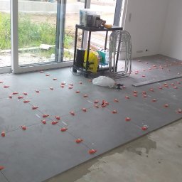 RAJ-BUD Usługi Ogólnobudowlane Robert Fujak - Szare płytki podłogowe ułożone w kwadraty z pomarańczowymi krzyżykami dystansowymi, widoczne narzędzia, wiadro i stolik z akcesoriami w jasnym, pustym pomieszczeniu z dużym oknem.