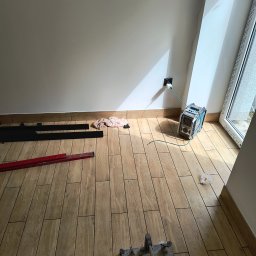 RAJ-BUD Usługi Ogólnobudowlane Robert Fujak - Ułożone panele podłogowe imitujące drewno w jasnym pomieszczeniu z widocznymi narzędziami: poziomica, listwy przypodłogowe i przecinarka do paneli, przy ścianie agregat prądotwórczy podłączony...