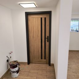 RAJ-BUD Usługi Ogólnobudowlane Robert Fujak - Wnętrze po remoncie: drewnopodobne płytki podłogowe, drewniane drzwi z czarnymi wstawkami i ościeżnicą, białe ściany z widocznymi gniazdkami, wiadro z zaprawą i farby w tle.