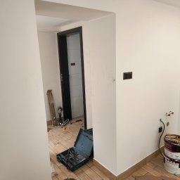 RAJ-BUD Usługi Ogólnobudowlane Robert Fujak - Wnętrze w trakcie prac wykończeniowych: widoczne narzędzia, puszki z farbą Magnat Ultra Matt, fragment świeżo pomalowanej ściany, drewnopodobna listwa przypodłogowa i płytki imitujące drewno.