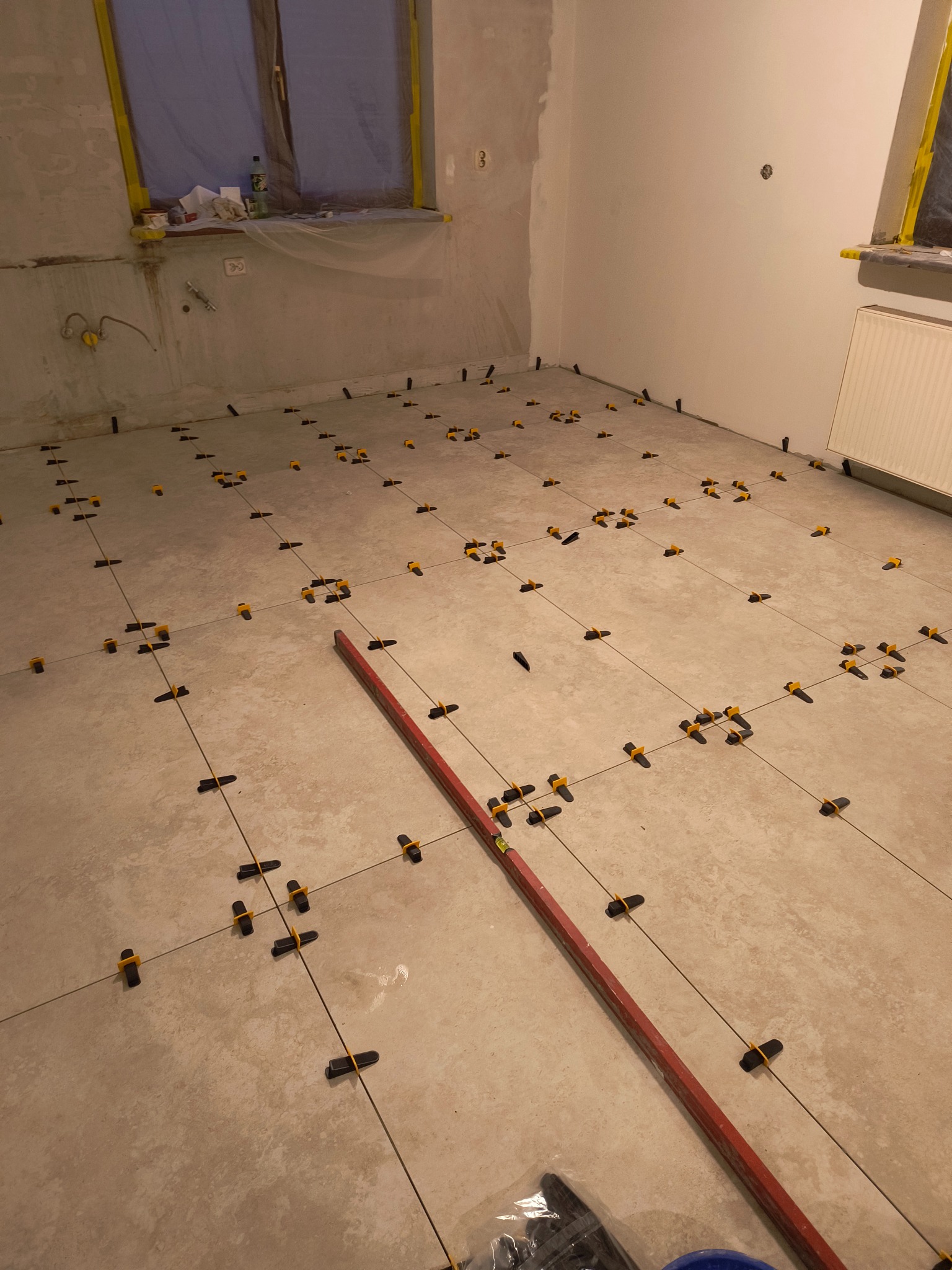 Układanie płytek podłogowych w jasnym kolorze z użyciem systemu poziomowania płytek, widoczna poziomica i wiadro, okno zabezpieczone folią malarską.