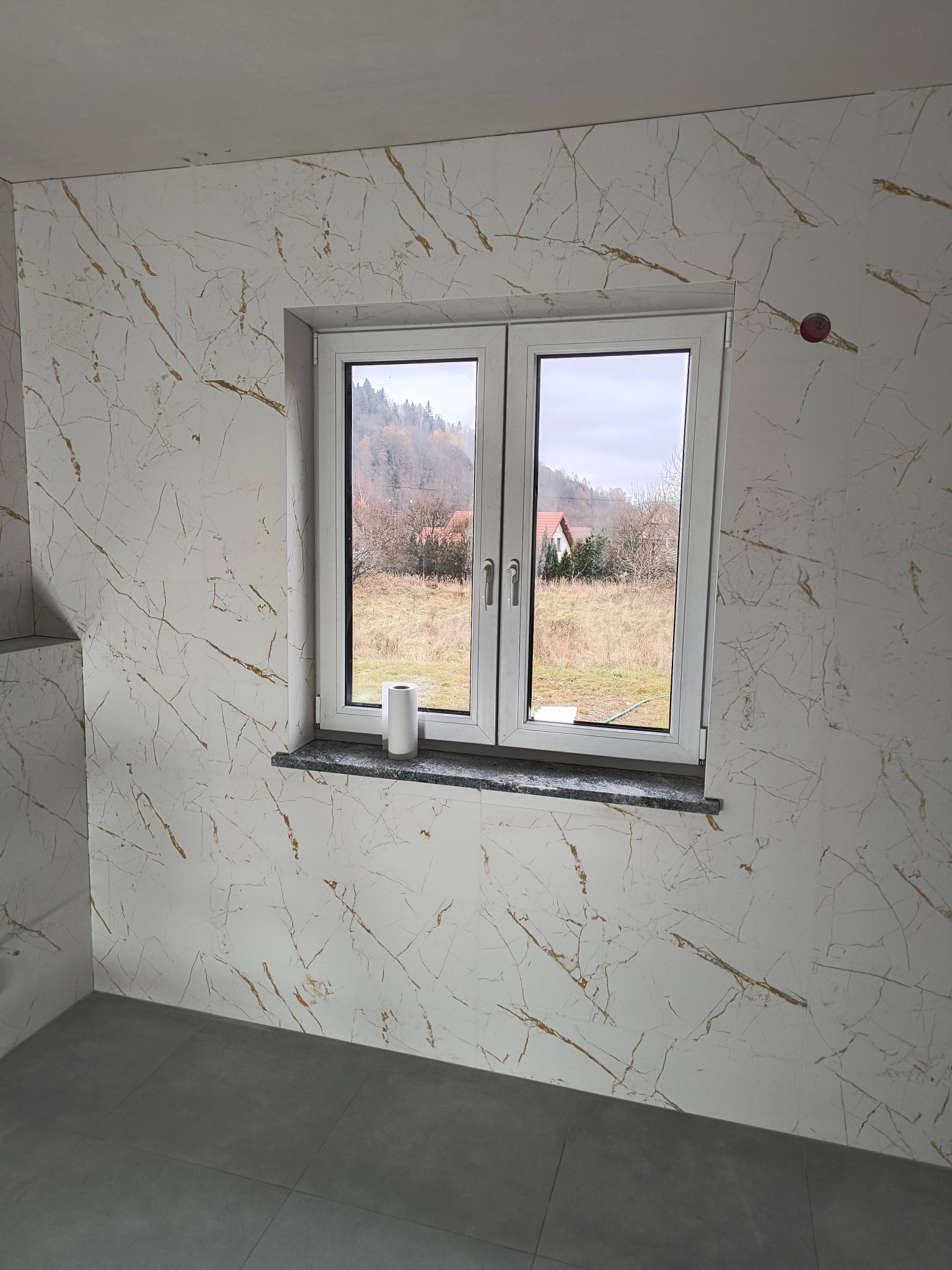 Wykończona łazienka z marmurowymi płytkami ściennymi w odcieniach bieli i złota, szare płytki podłogowe, białe okno z widokiem na zewnątrz i rolka papieru na parapecie.