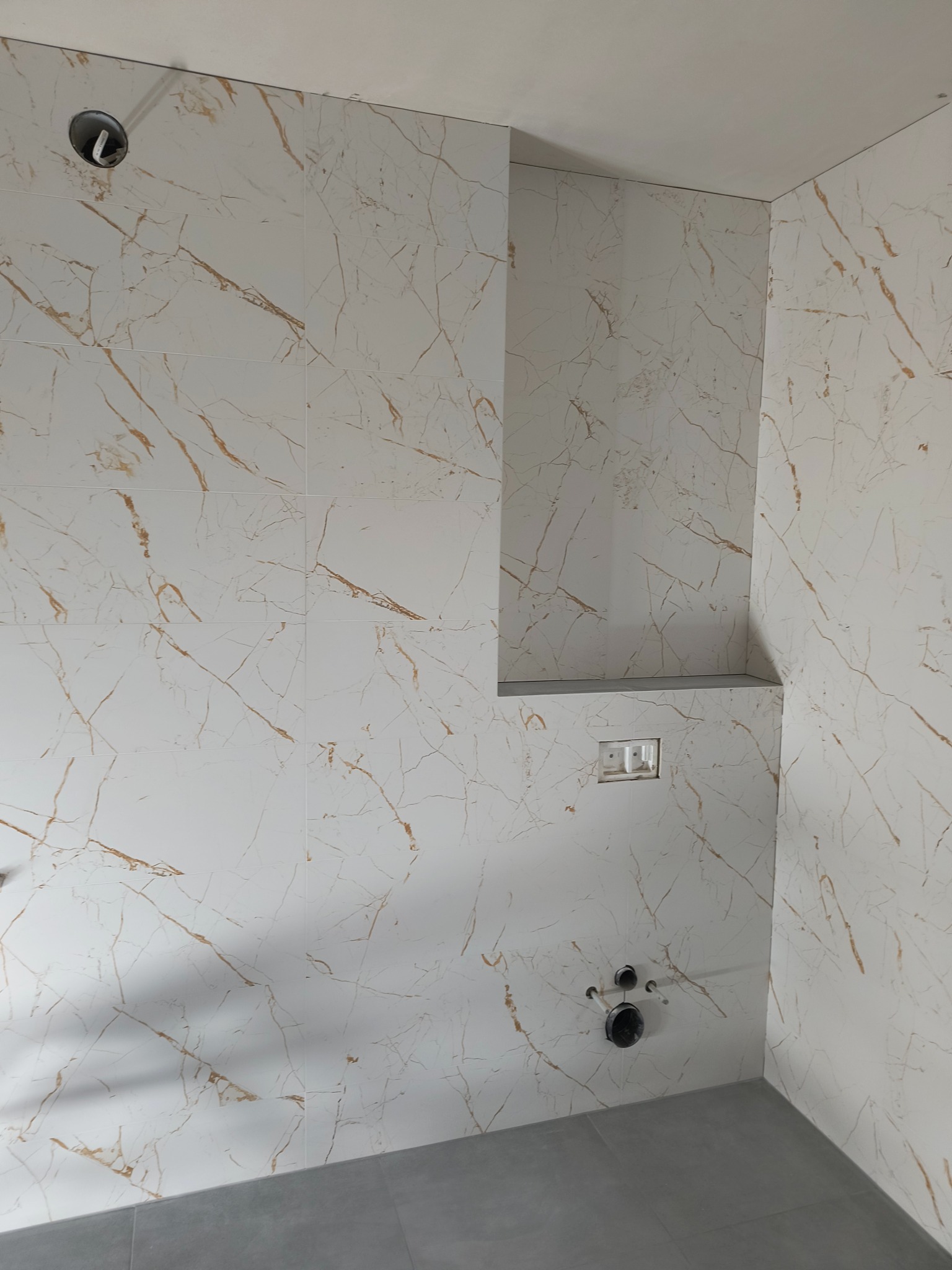 Wnętrze łazienki w trakcie wykańczania, widoczne białe płytki imitujące marmur z złotymi żyłami na ścianach, szara podłoga, przygotowane instalacje wodno-kanalizacyjne i elektryczne, wnęka...