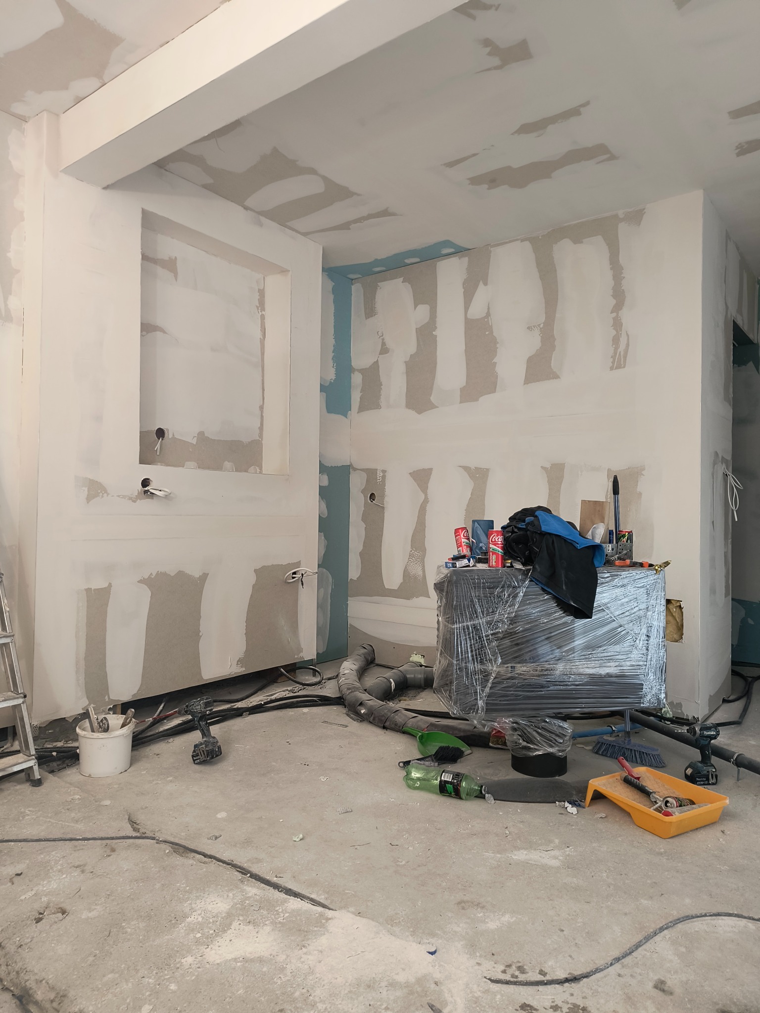 Wnętrze w trakcie prac wykończeniowych: ściany i sufit pokryte gładzią, widoczne przewody elektryczne, narzędzia malarskie i meble zabezpieczone folią.