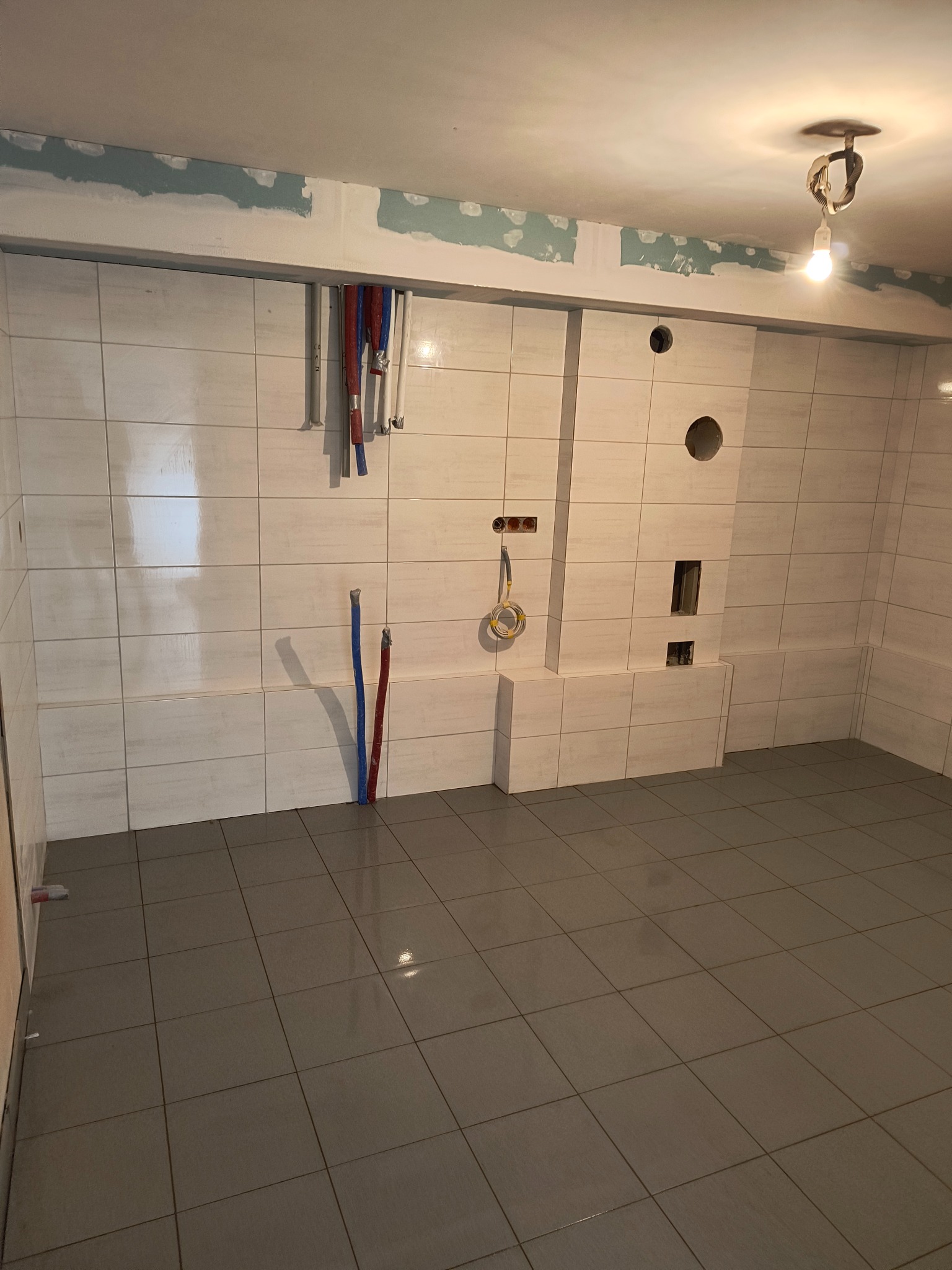 Wnętrze łazienki w trakcie remontu, widoczne białe płytki ścienne z szarą fugą, szare płytki podłogowe, wystające rury instalacji wodnej i elektrycznej, oraz fragment ściany z nałożoną gładzią...