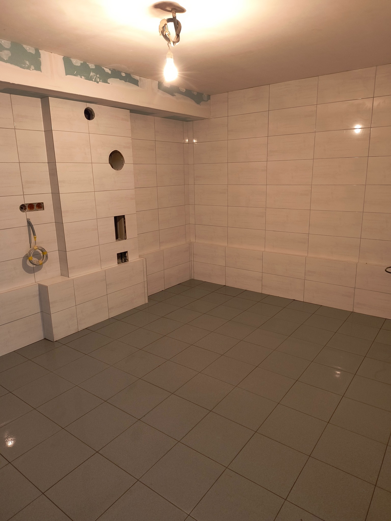 Wnętrze łazienki w trakcie wykańczania: ułożone szare płytki podłogowe i jasne płytki ścienne, widoczne otwory na instalacje, surowy sufit z widocznymi śladami prac przygotowawczych...