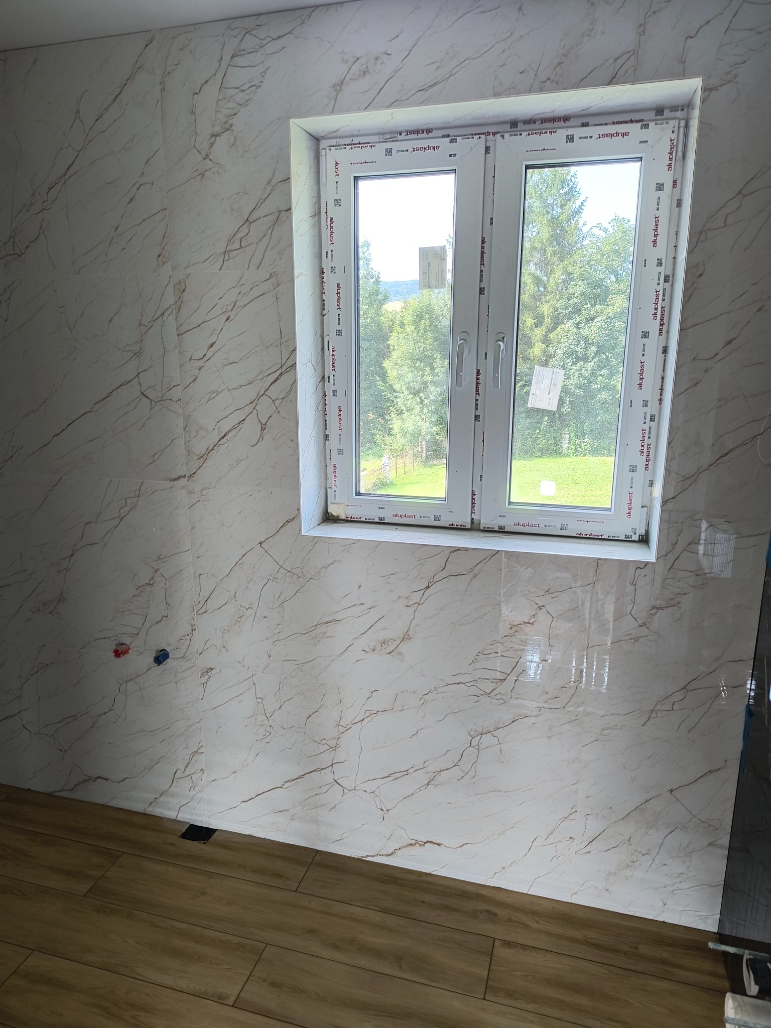 Wykończona łazienka z marmurowymi płytkami na ścianach, widok na okno z widokiem na zieleń, drewnopodobne panele na podłodze, przygotowane przyłącza wodne.