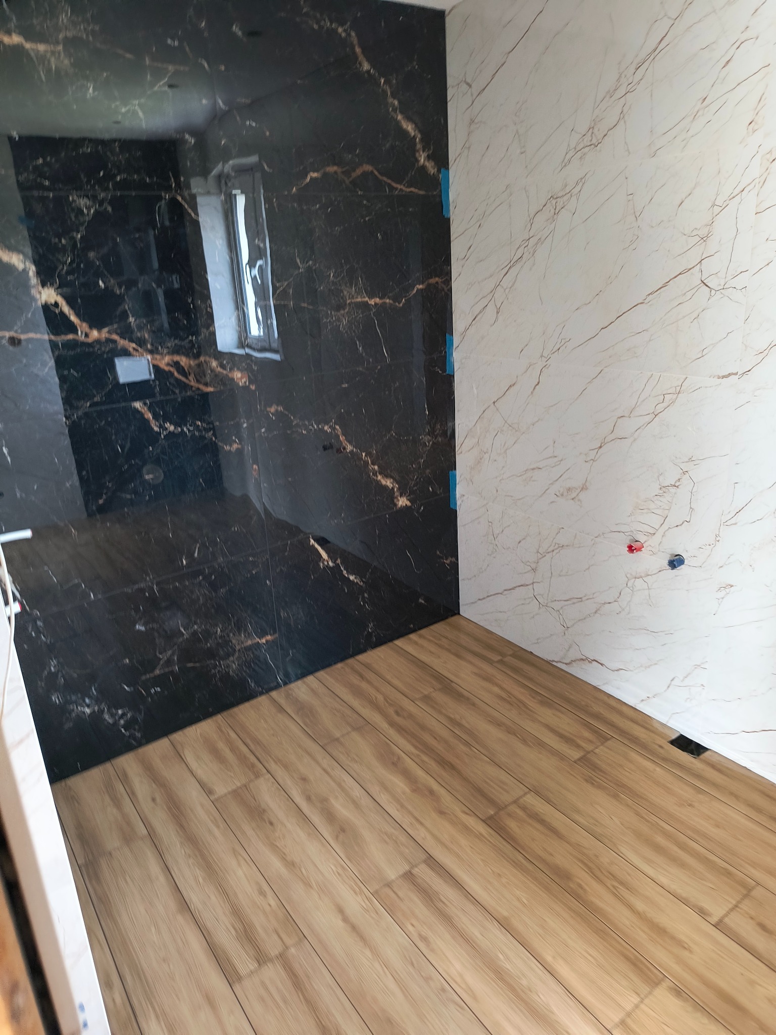 Wykończona łazienka z czarnymi i białymi marmurkowymi płytkami oraz panelami podłogowymi imitującymi drewno, widoczne niebieskie taśmy malarskie zabezpieczające krawędzie.