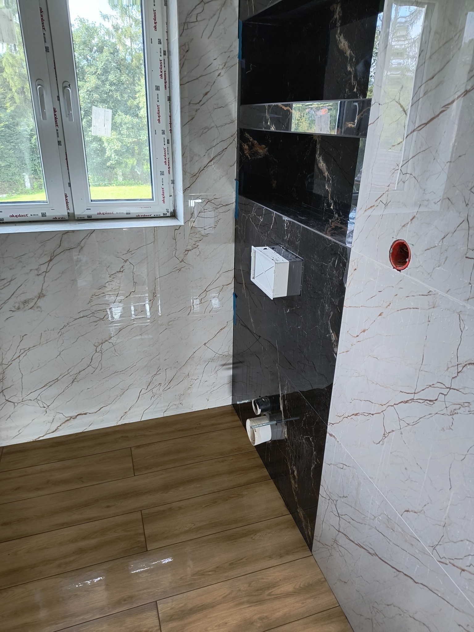 Wykończona łazienka z marmurowymi płytkami w odcieniach bieli i czerni, drewnopodobna podłoga, okno z widokiem na zieleń, zabudowana półka z czarnego marmuru z widocznymi elementami instalacji.