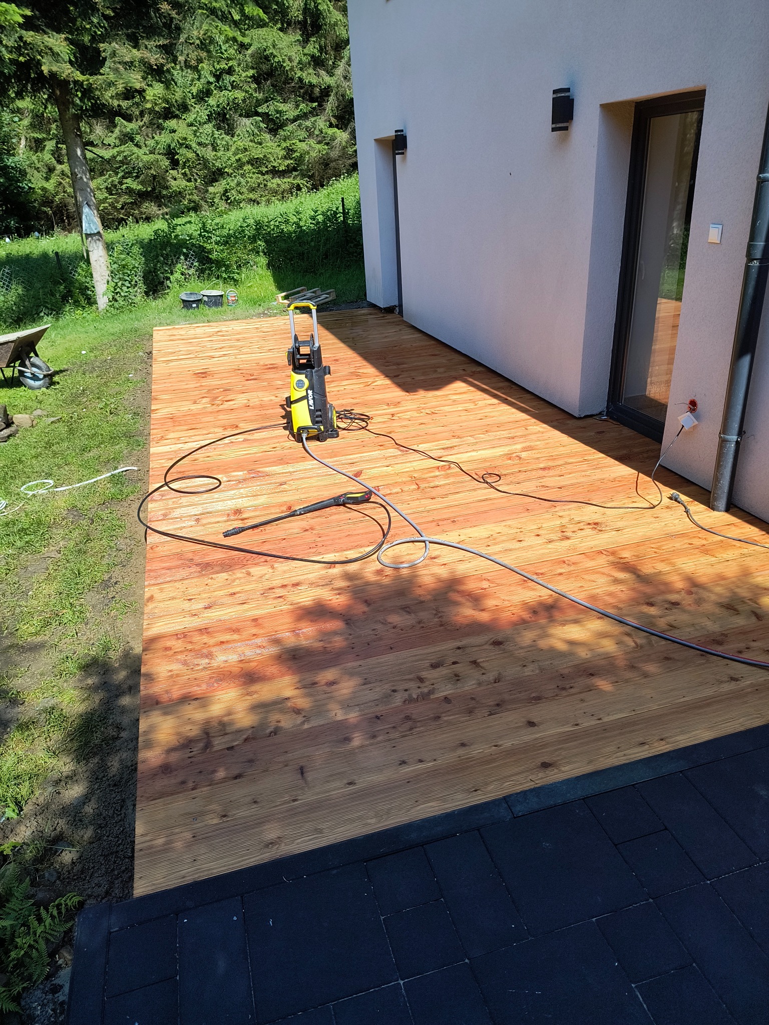 Czyszczenie drewnianego tarasu przy domu myjką ciśnieniową, widoczny fragment ściany budynku, trawnik i drzewa w tle.