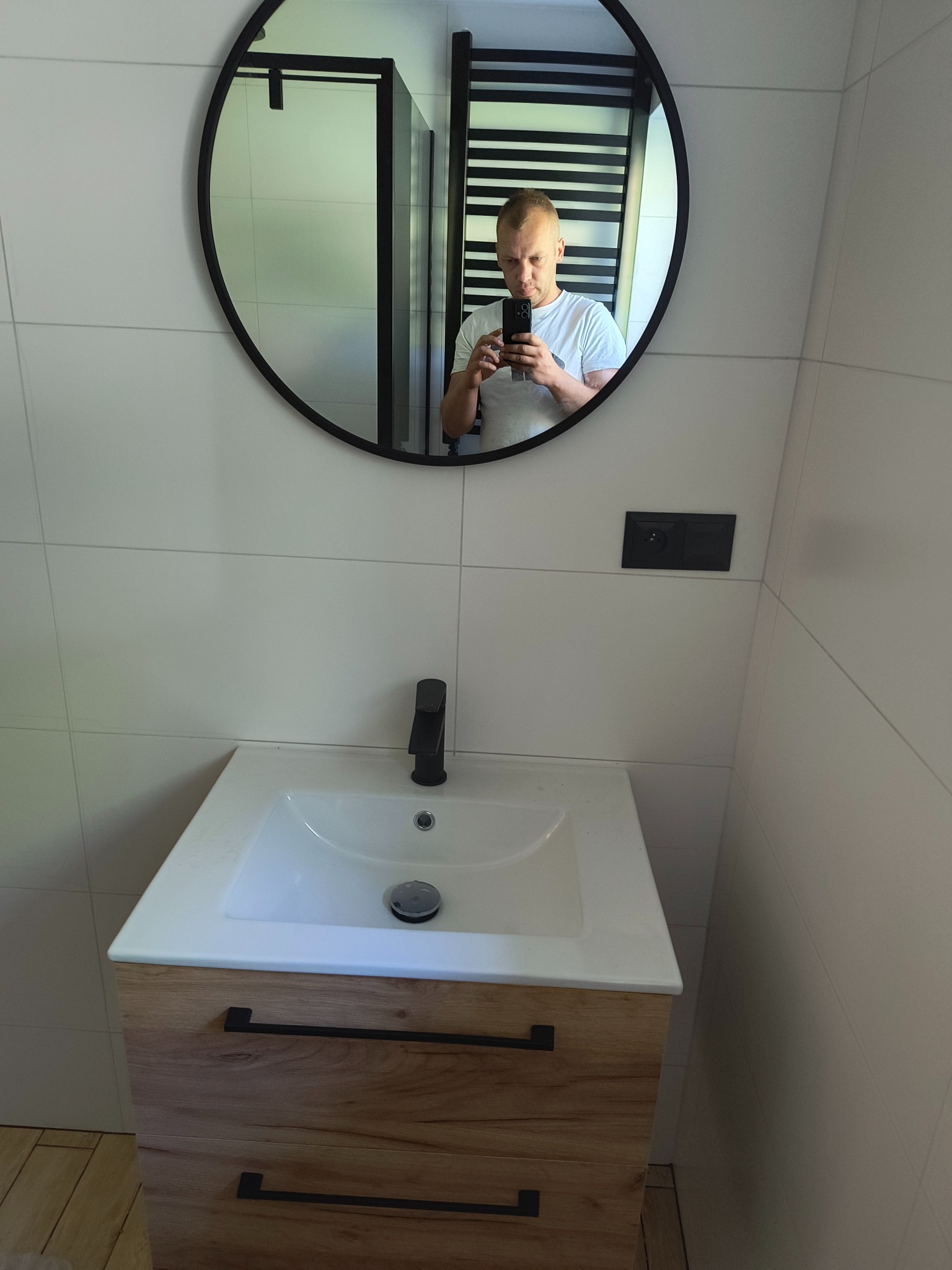 Nowoczesna łazienka z białą umywalką nablatową na szafce z drewnianym fornirem i czarnymi uchwytami, czarna bateria umywalkowa, białe płytki na ścianach i okrągłe lustro z czarną ramą, w którym...