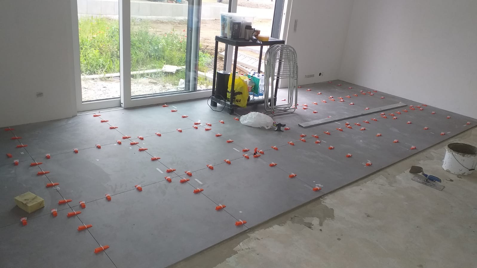 Szare płytki podłogowe ułożone w kwadraty z pomarańczowymi krzyżykami dystansowymi, widoczne narzędzia, wiadro i stolik z akcesoriami w jasnym, pustym pomieszczeniu z dużym oknem.