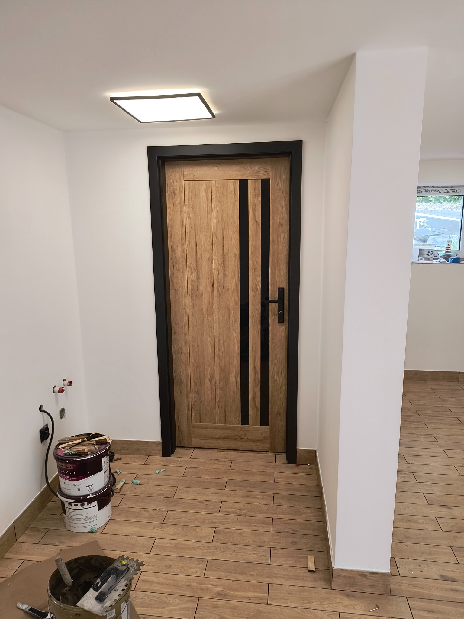 Wnętrze po remoncie: drewnopodobne płytki podłogowe, drewniane drzwi z czarnymi wstawkami i ościeżnicą, białe ściany z widocznymi gniazdkami, wiadro z zaprawą i farby w tle.