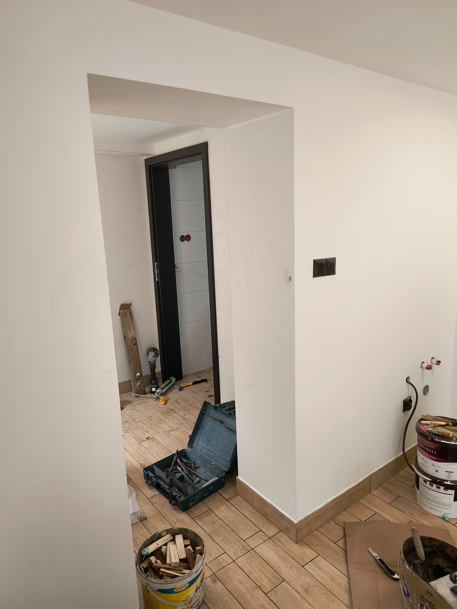 Wnętrze w trakcie prac wykończeniowych: widoczne narzędzia, puszki z farbą Magnat Ultra Matt, fragment świeżo pomalowanej ściany, drewnopodobna listwa przypodłogowa i płytki imitujące drewno.