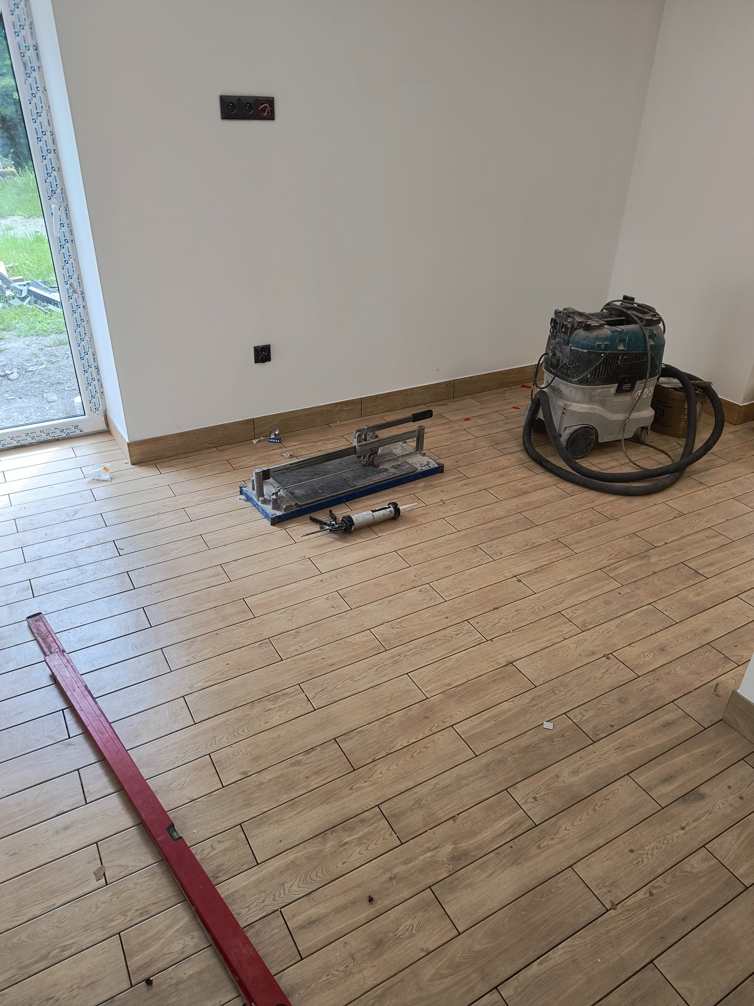 Układanie drewnopodobnych płytek podłogowych w jasnym pomieszczeniu, widoczny poziomica, przecinarka do płytek, odkurzacz przemysłowy oraz gniazdka elektryczne.