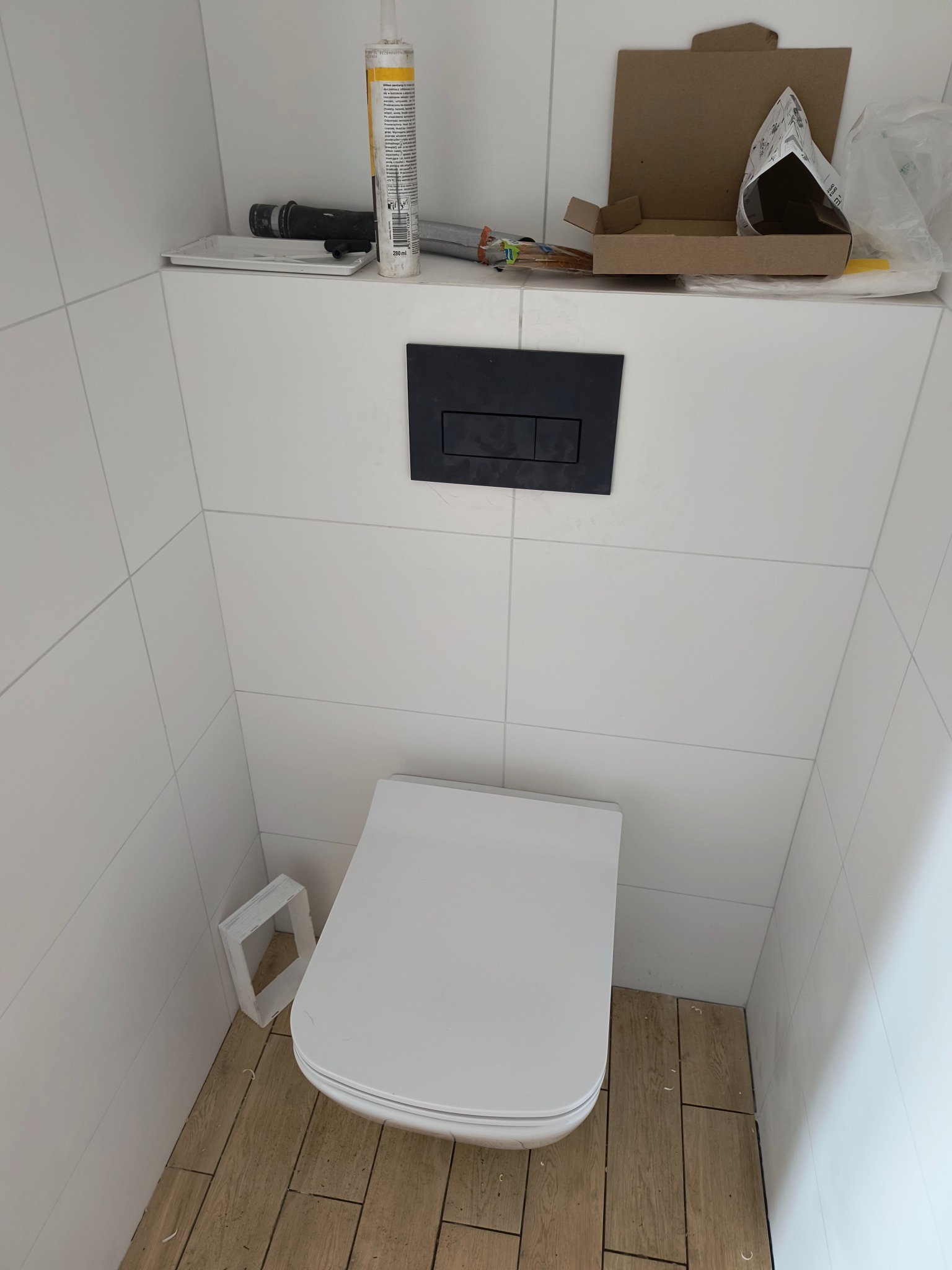 Nowoczesna toaleta podwieszana z czarnym przyciskiem spłukującym, białe płytki ścienne i drewnopodobne płytki podłogowe, widoczne akcesoria montażowe i opakowania na półce nad toaletą.