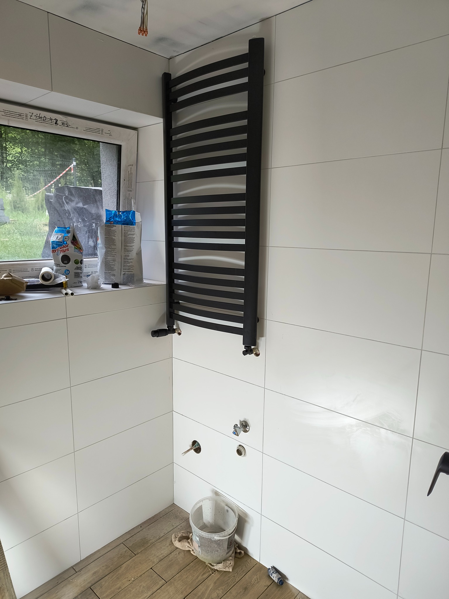 Łazienka w trakcie wykańczania: białe płytki ścienne, drewnopodobne płytki podłogowe, czarny grzejnik drabinkowy, worki z zaprawą na parapecie okna, widoczne przyłącza wodne bez armatury.