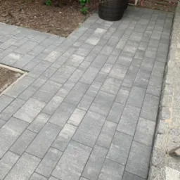 Ułożona szara kostka brukowa tworząca ścieżkę w ogrodzie z wiklinowym fotelem w tle.