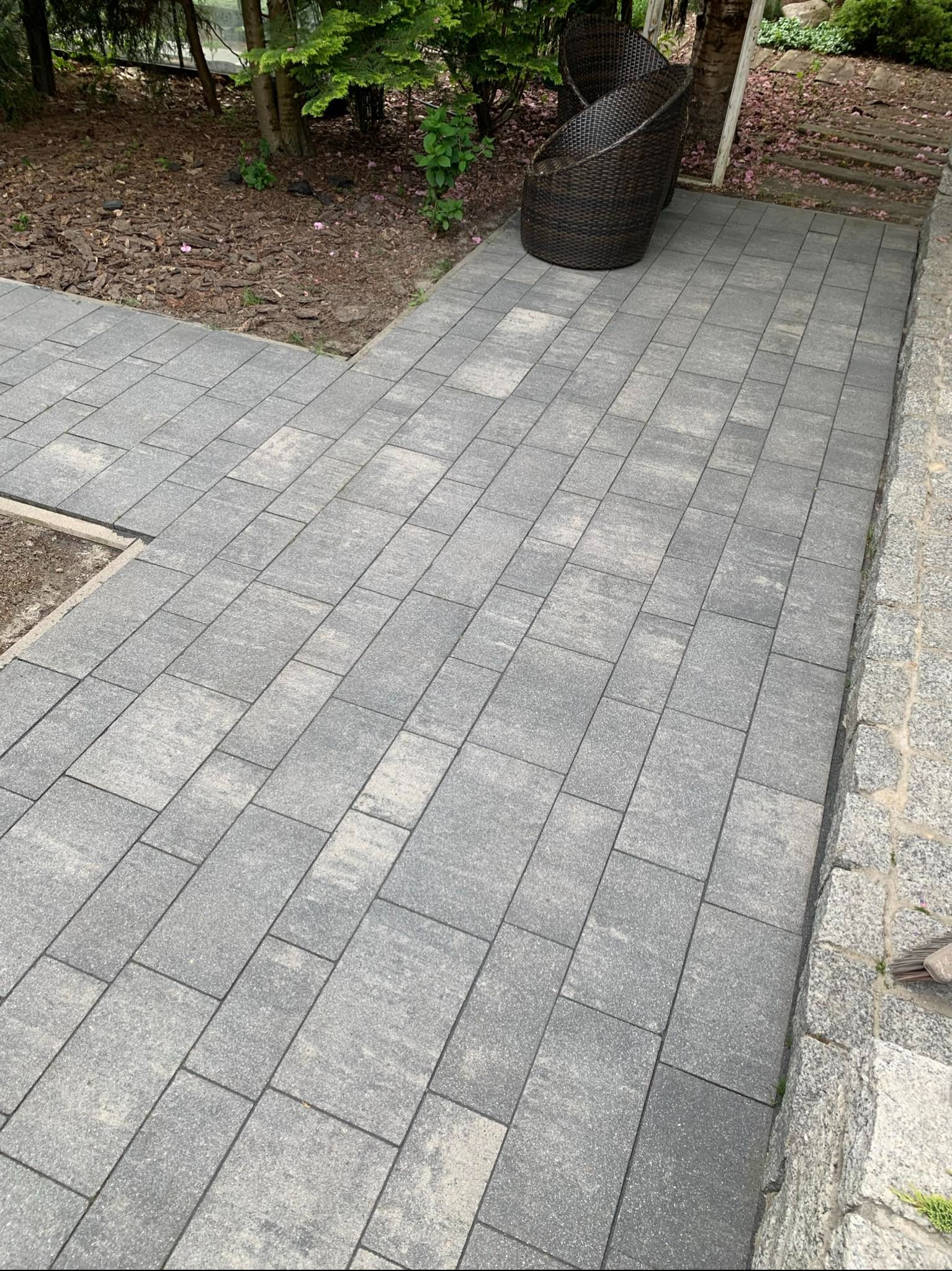 Ułożona szara kostka brukowa tworząca ścieżkę w ogrodzie z wiklinowym fotelem w tle.