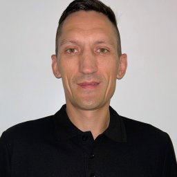 FIRMA DEA NATALIIA HRUZKA - Portret mężczyzny w czarnej koszulce polo z logo Stanley Black&Decker na tle białej ściany. Uśmiechnięty, z krótkimi włosami i brązowymi oczami.