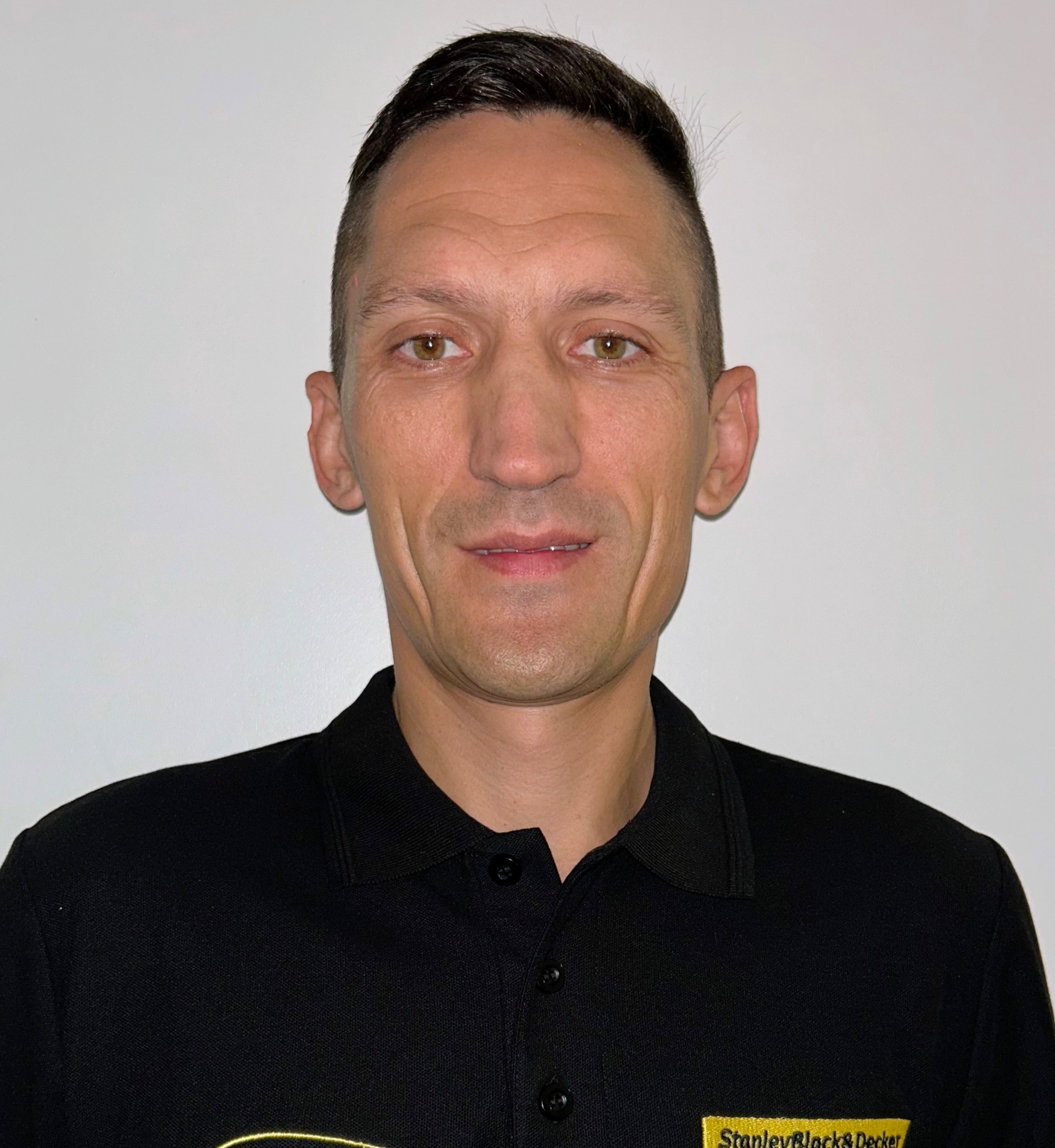 Portret mężczyzny w czarnej koszulce polo z logo Stanley Black&Decker na tle białej ściany. Uśmiechnięty, z krótkimi włosami i brązowymi oczami.