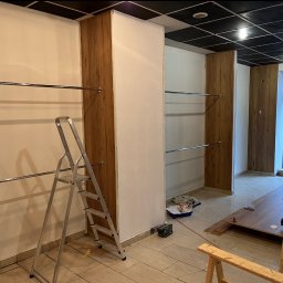 ART STUDIO - Wnętrze lokalu komercyjnego w trakcie wykańczania: pomalowane na biało ściany, drewniane panele ścienne, metalowe relingi na ubrania, drabina, kuweta malarska i narzędzia malarskie na podłodze...