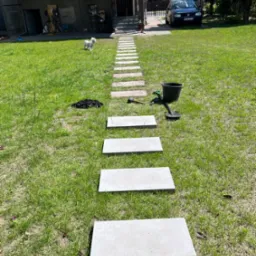 Ścieżka z betonowych płyt ułożona w trawie, narzędzia ogrodnicze obok, mały biały pies w tle, dom i samochód w oddali.