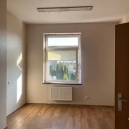 Pusty pokój z drewnianą podłogą, beżowymi ścianami i oknem z widokiem na zieleń. Grzejnik pod oknem. Otwarta drewniana ościeżnica drzwi po prawej stronie kadru.