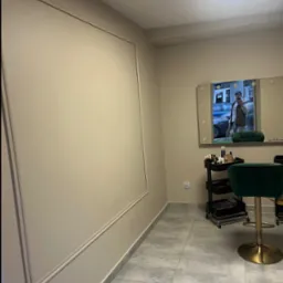 Ściana w salonie kosmetycznym po malowaniu, z dekoracyjną ramą. W tle lustro z oświetleniem i zielone krzesło. Widoczny wózek z kosmetykami i szara podłoga.