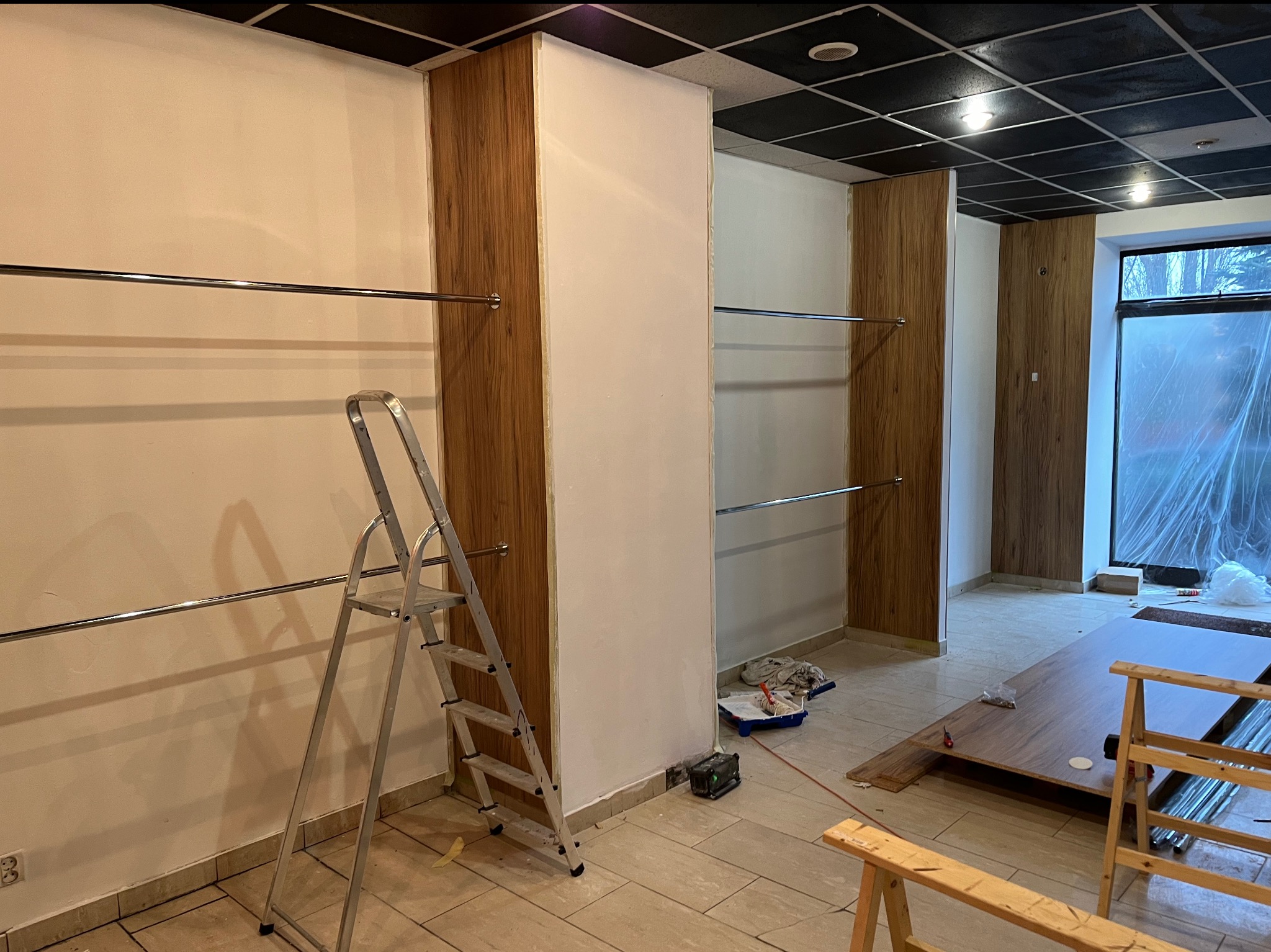 Wnętrze lokalu komercyjnego w trakcie wykańczania: pomalowane na biało ściany, drewniane panele ścienne, metalowe relingi na ubrania, drabina, kuweta malarska i narzędzia malarskie na podłodze...