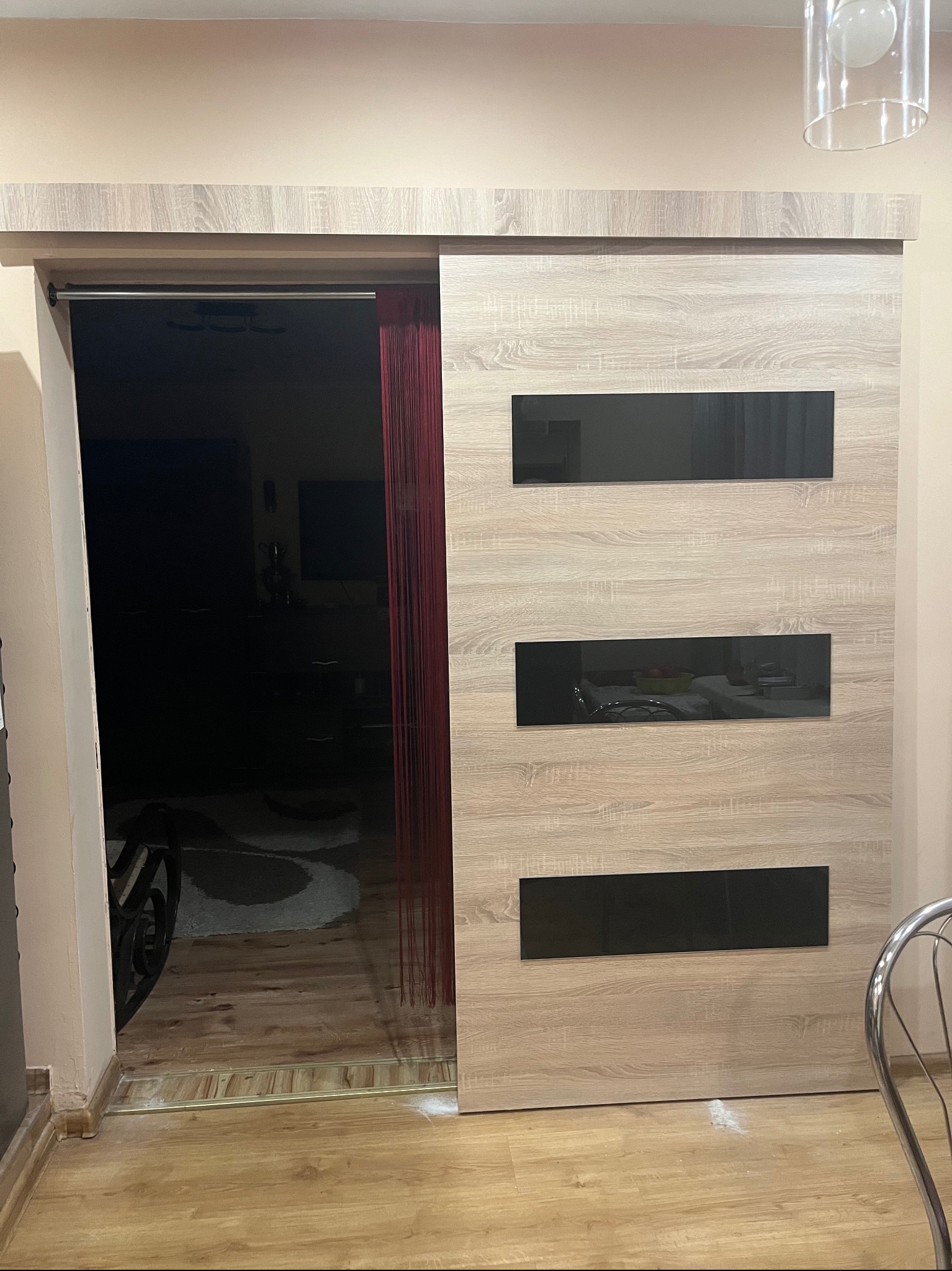 Drewniane drzwi przesuwne z trzema poziomymi, czarnymi szybami, częściowo otwarte, z widocznymi czerwonymi sznurkami dekoracyjnymi i fragmentem wnętrza pokoju.