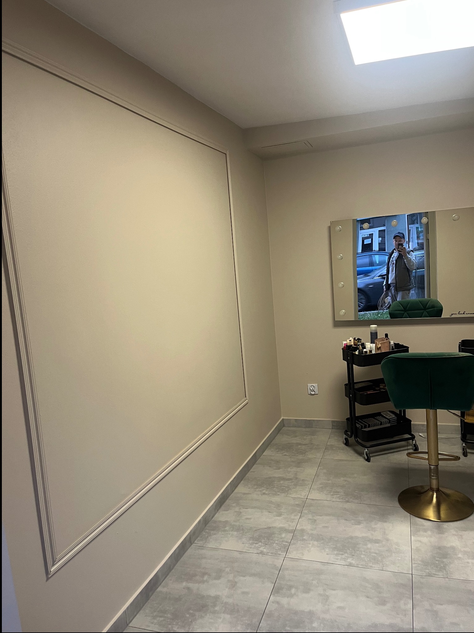 Ściana w salonie kosmetycznym po malowaniu, z dekoracyjną ramą. W tle lustro z oświetleniem i zielone krzesło. Widoczny wózek z kosmetykami i szara podłoga.