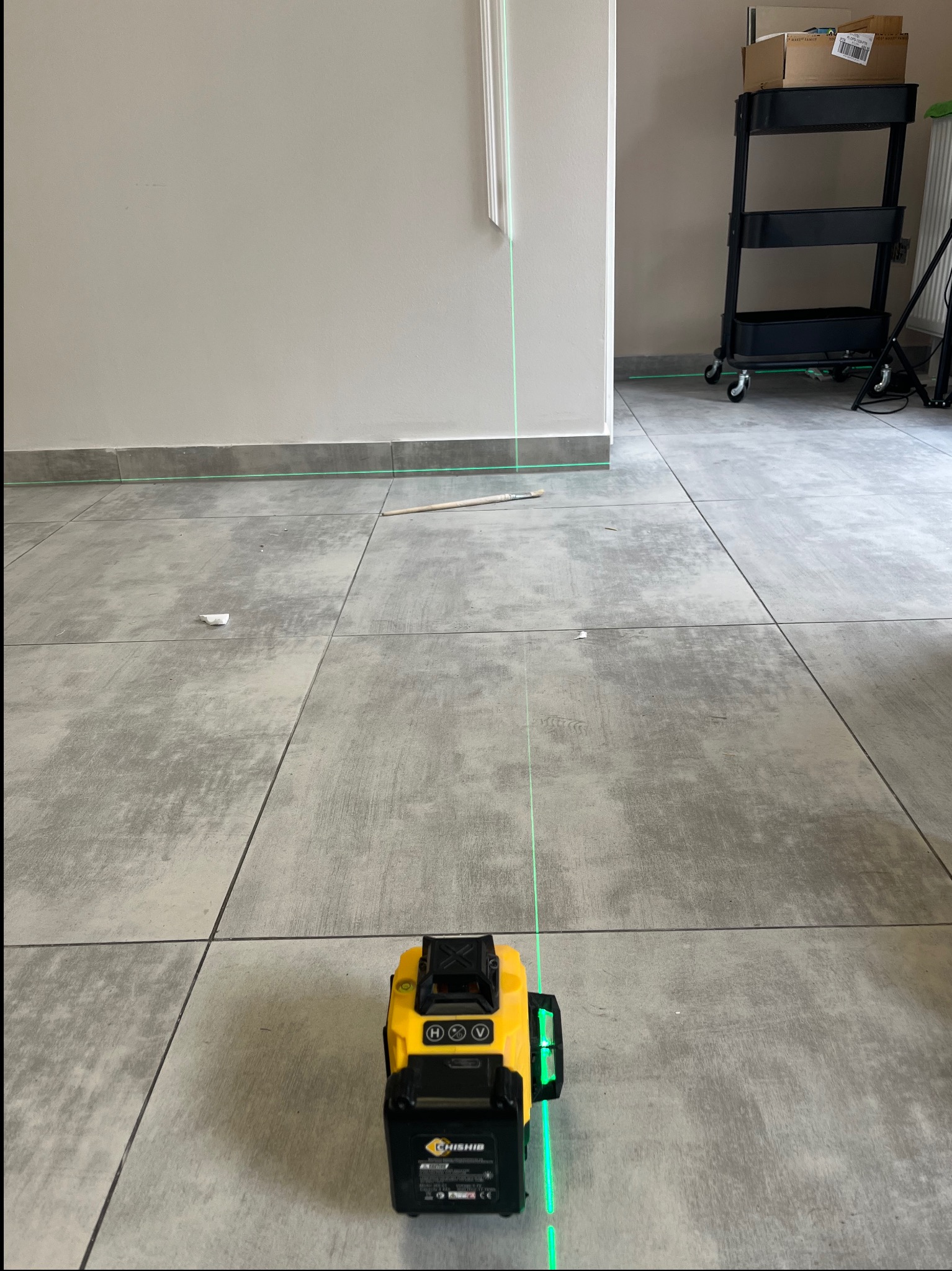 Laser krzyżowy wyświetla zielone linie na szarych płytkach podłogowych w jasnym pomieszczeniu. Widoczny fragment ściany i wózek z półkami w tle.