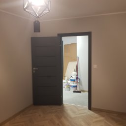 Maksym Pavlishchuk - Pokój w trakcie remontu: widoczna podłoga ułożona w jodełkę, nowoczesna lampa wisząca i otwarte drzwi do pomieszczenia z puszkami farb i narzędziami malarskimi.