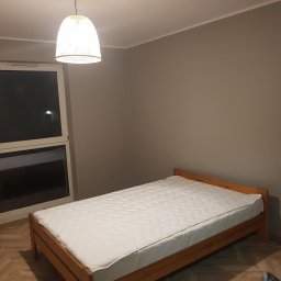 Maksym Pavlishchuk - Pokój z drewnianym łóżkiem z materacem, lampą wiszącą i widokiem przez okno, świeżo po remoncie.