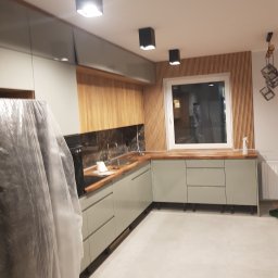 Maksym Pavlishchuk - Nowoczesna kuchnia w trakcie wykańczania: szare szafki z drewnianym blatem, drewniana ściana z lamelami, czarne lampy sufitowe, sprzęt AGD częściowo zakryty folią ochronną.