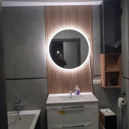 Maksym Pavlishchuk - Nowoczesna łazienka z okrągłym lustrem podświetlanym LED, umywalką z szafką w folii ochronnej, drewnianymi panelami dekoracyjnymi i szarą płytką.