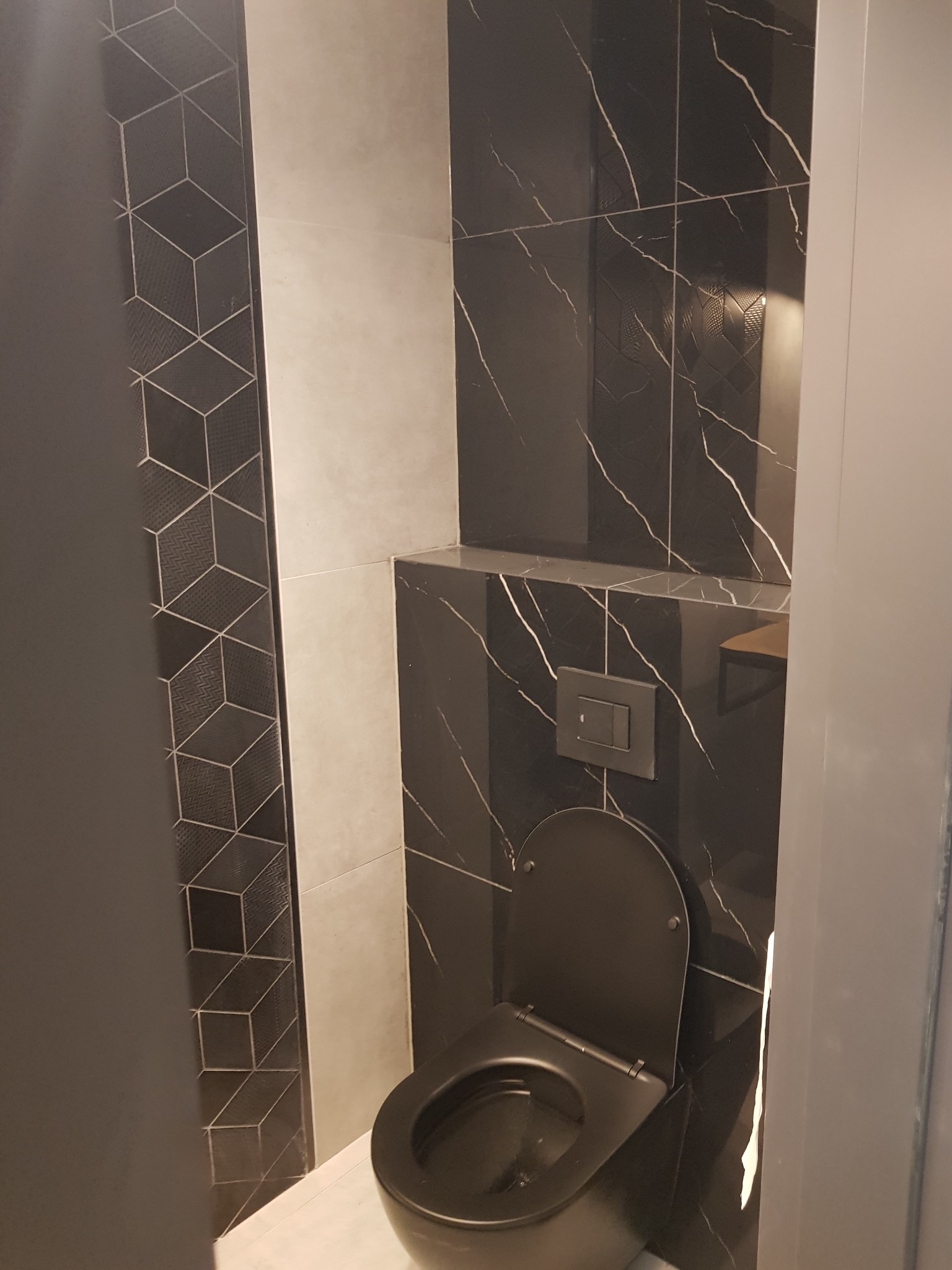 Nowoczesna toaleta w łazience z czarno-białymi płytkami: marmurkowe na ścianie za toaletą i geometryczne na ścianie po lewej stronie, widok zza futryny drzwi.