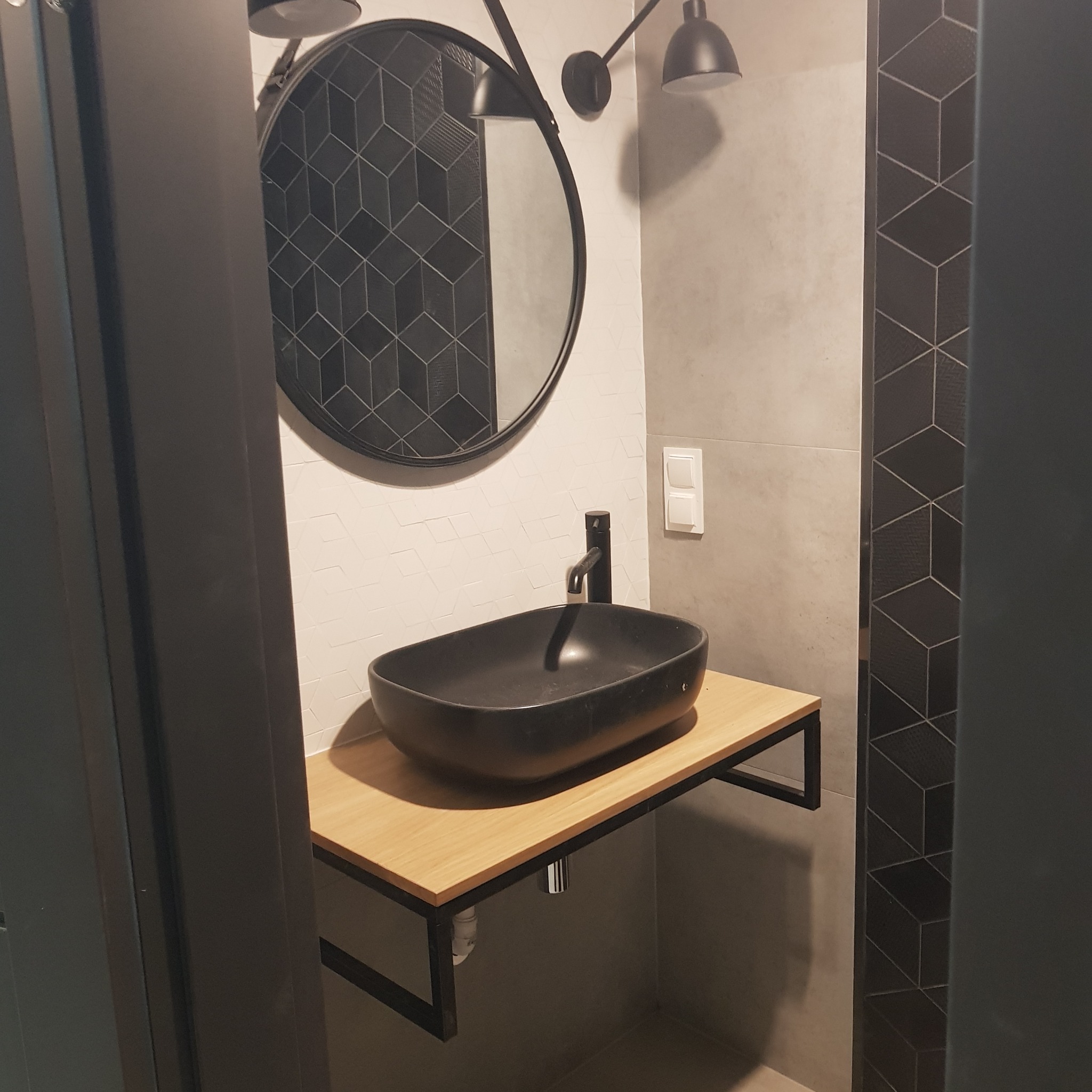 Nowoczesna łazienka z czarną, owalną umywalką nablatową na drewnianym blacie z metalowym stelażem, okrągłe lustro z podwójnym oświetleniem punktowym, szara ściana i dekoracyjna płytka...