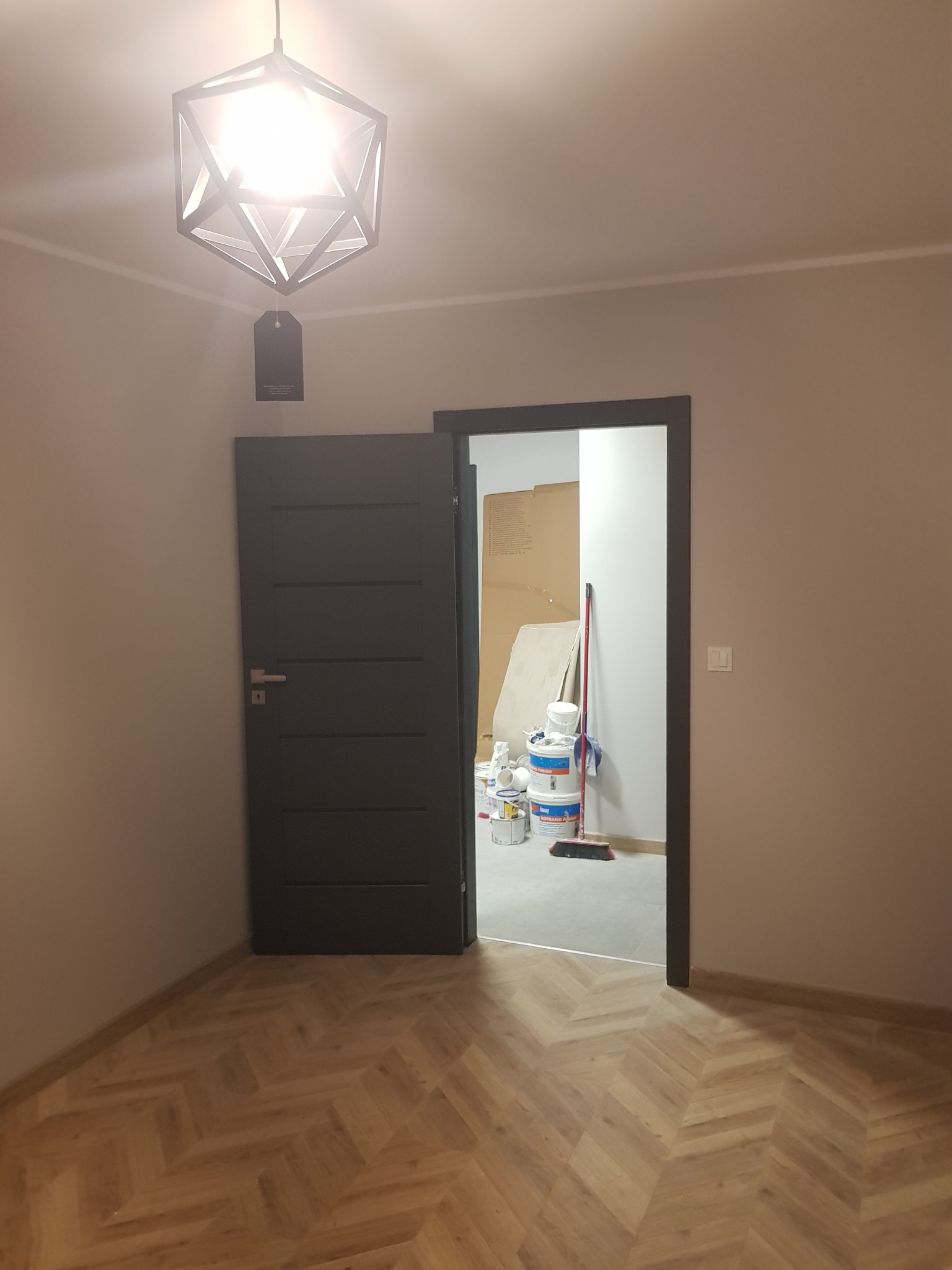 Pokój w trakcie remontu: widoczna podłoga ułożona w jodełkę, nowoczesna lampa wisząca i otwarte drzwi do pomieszczenia z puszkami farb i narzędziami malarskimi.