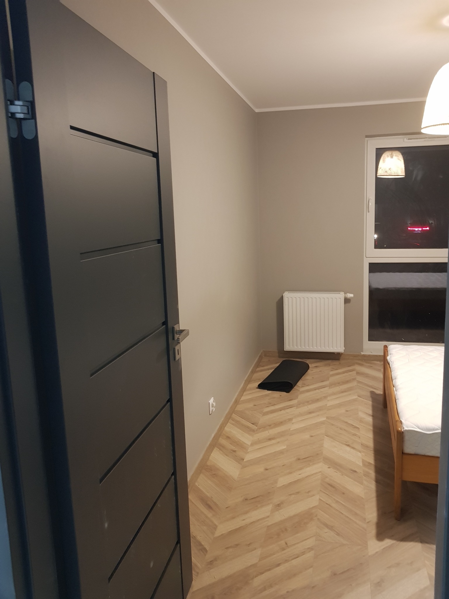 Ukończone układanie parkietu w jodełkę francuską, widoczny fragment pokoju z łóżkiem, grzejnikiem i czarną matą do ćwiczeń, świeżo po remoncie.