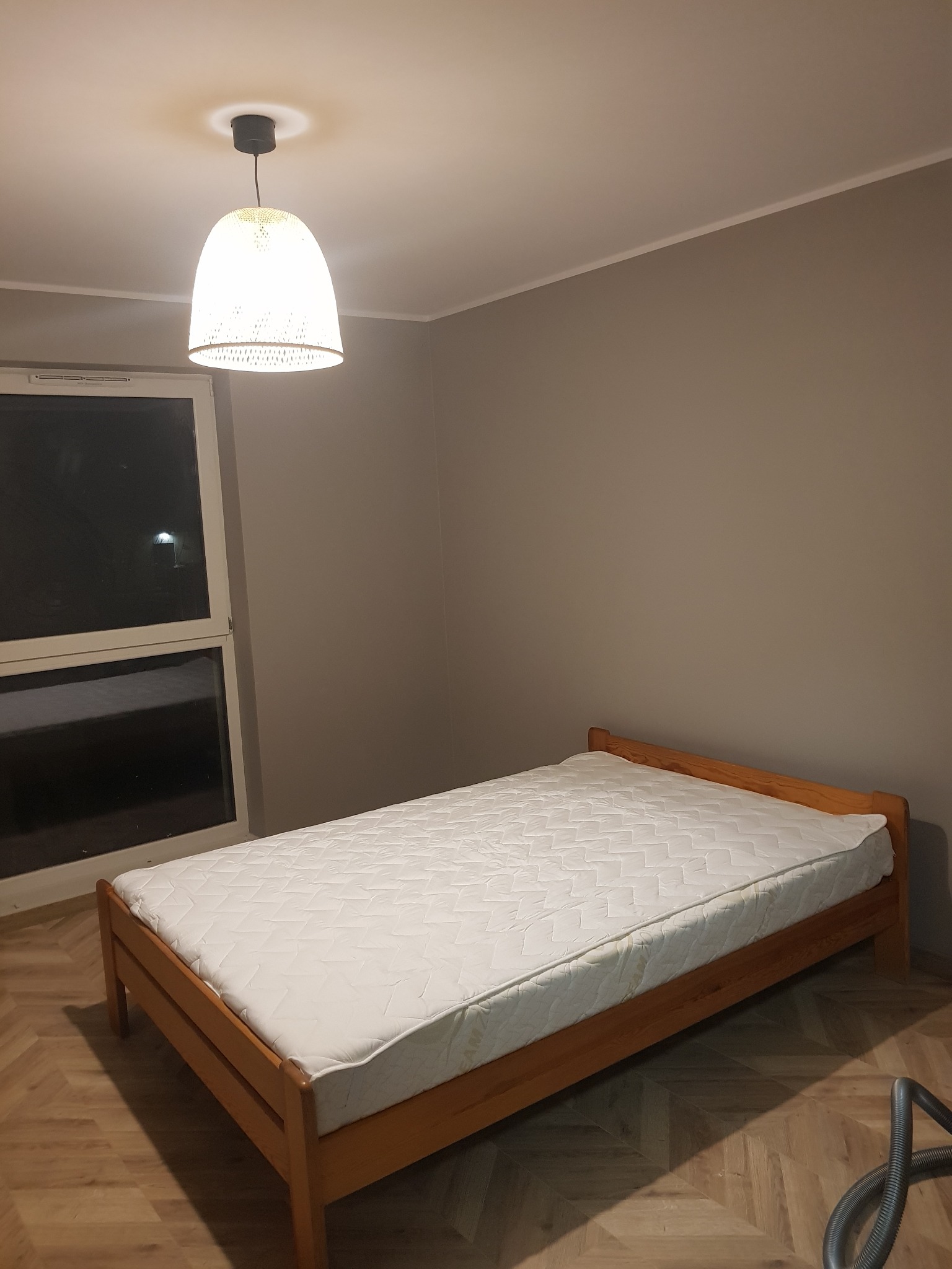 Pokój z drewnianym łóżkiem z materacem, lampą wiszącą i widokiem przez okno, świeżo po remoncie.