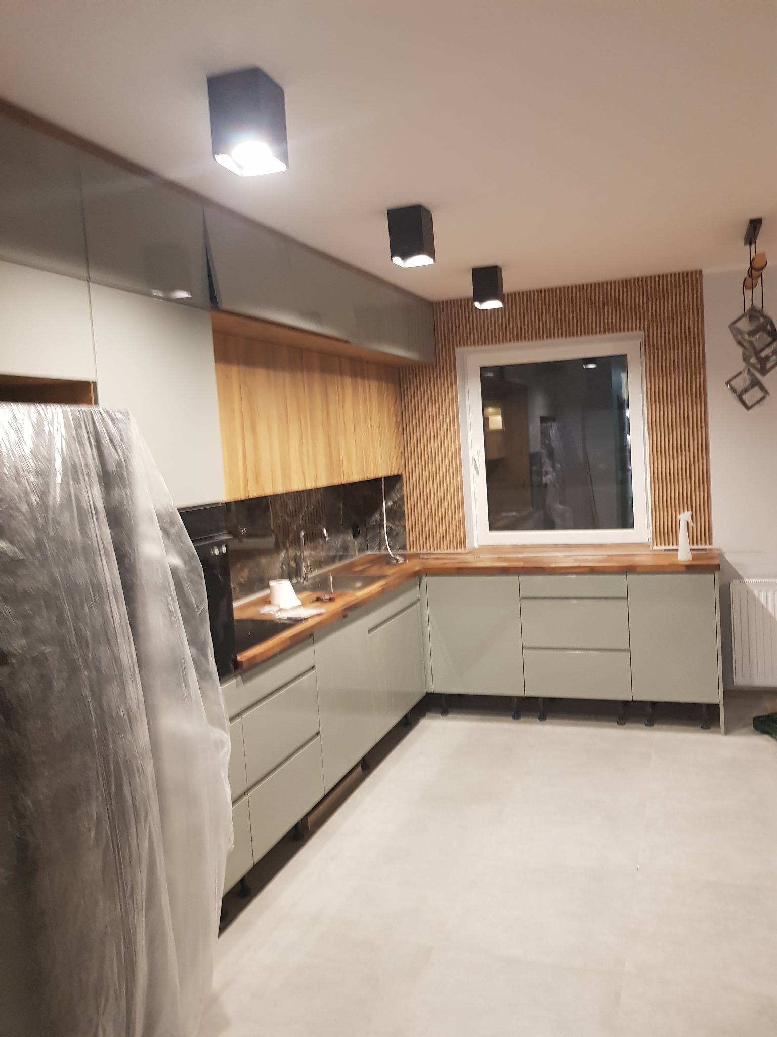 Nowoczesna kuchnia w trakcie wykańczania: szare szafki z drewnianym blatem, drewniana ściana z lamelami, czarne lampy sufitowe, sprzęt AGD częściowo zakryty folią ochronną.