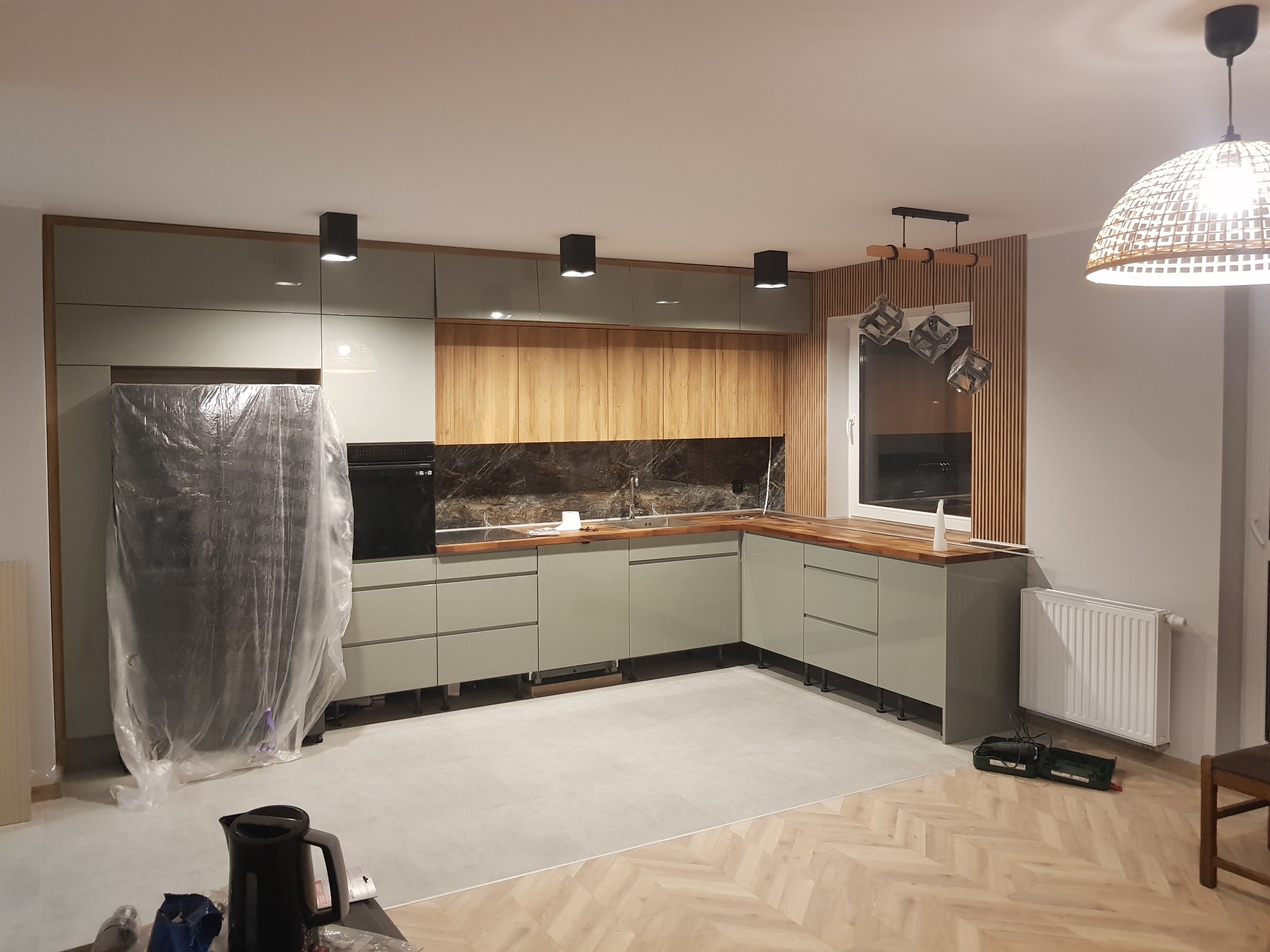 Nowoczesna kuchnia z szaro-zielonymi szafkami, drewnianym blatem i marmurowym panelem nad blatem, na podłodze parkiet ułożony w jodełkę, lodówka zabezpieczona folią, widoczne narzędzia i czajnik...